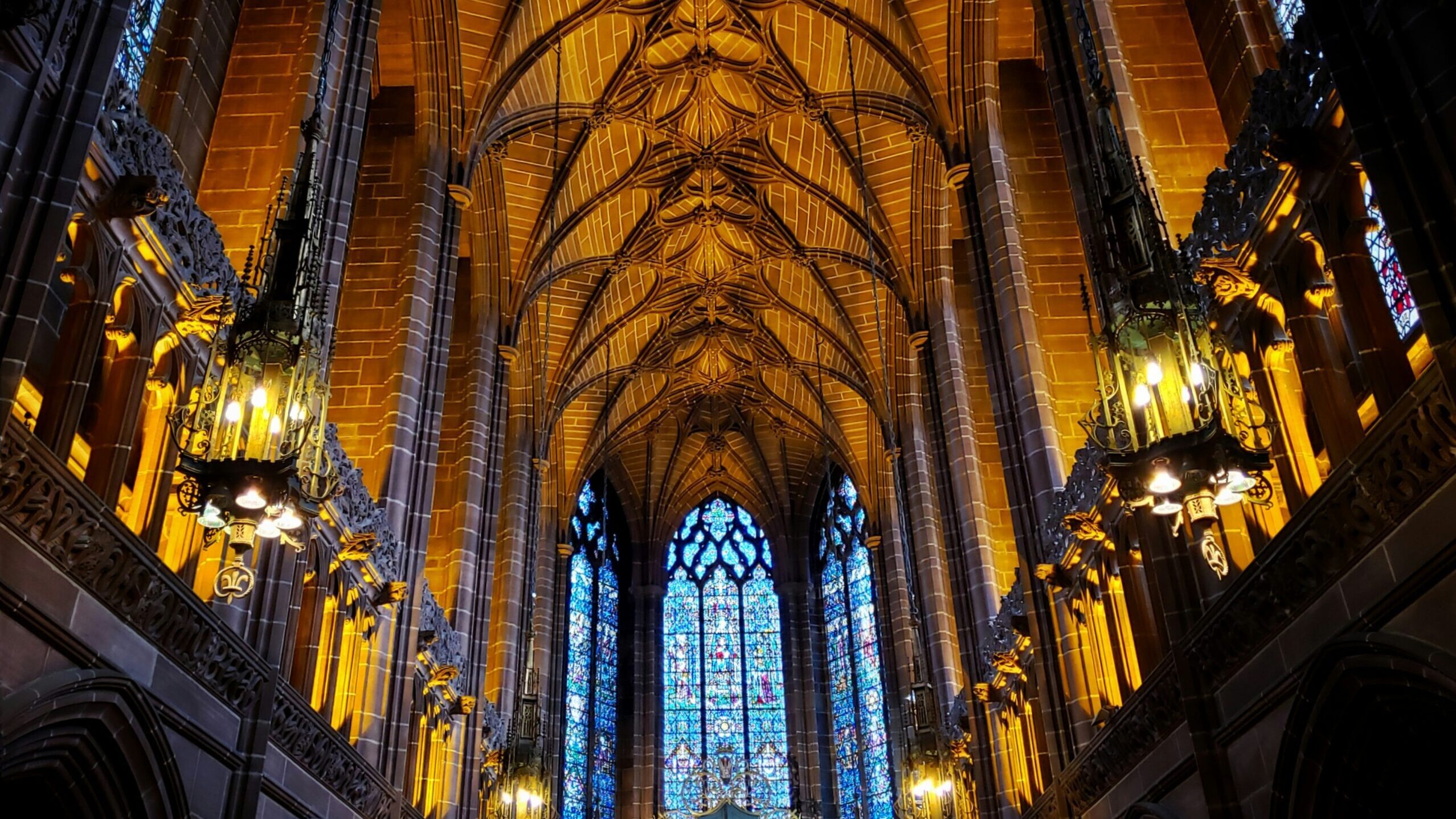 Stunning interior of a cathedral with ornate vaulted ceiling, glowing chandeliers, and large stained glass window.