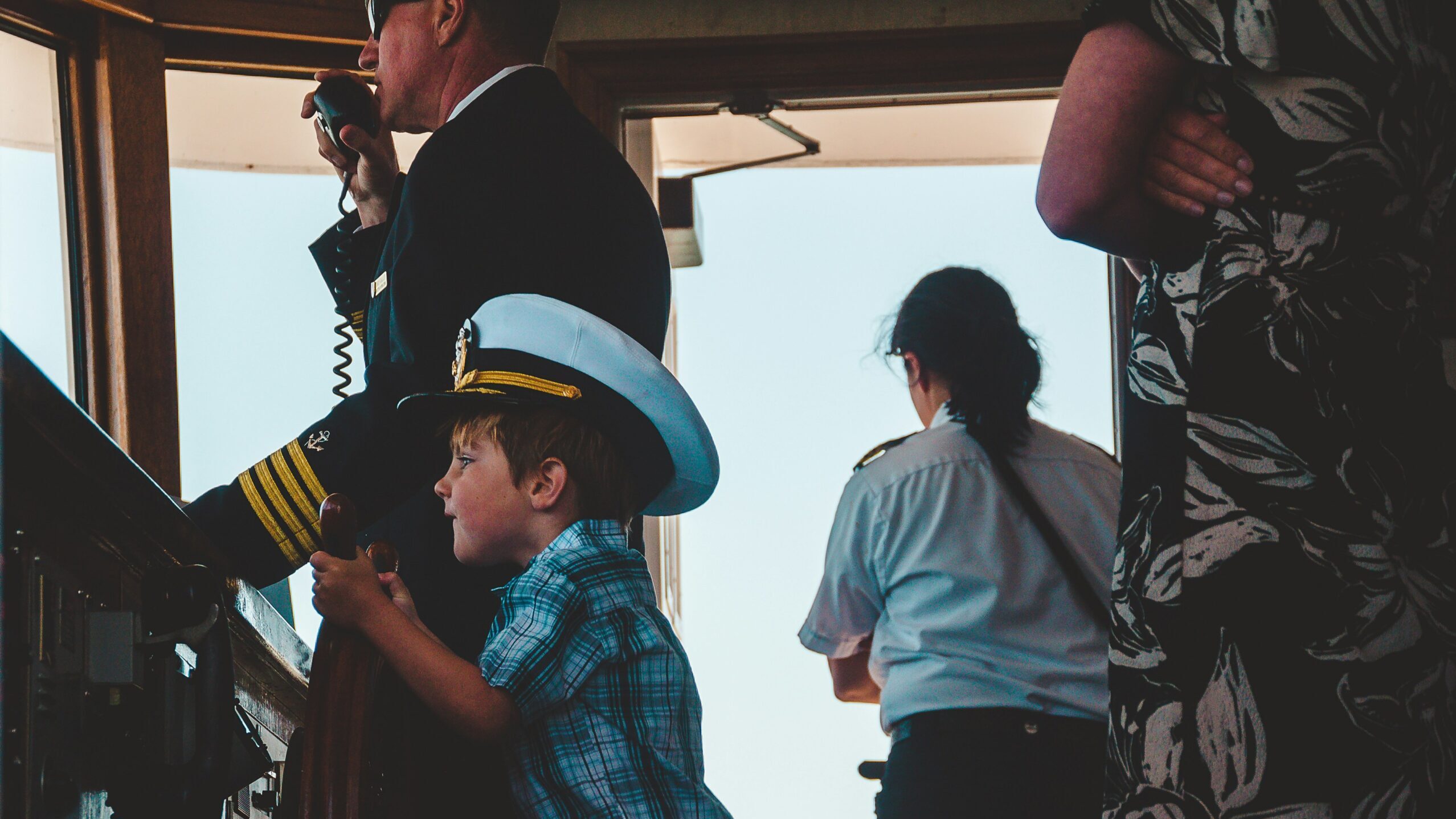 Young boy wearing a captain’s hat playfully steering a ship’s wheel alongside a uniformed officer on the bridge.