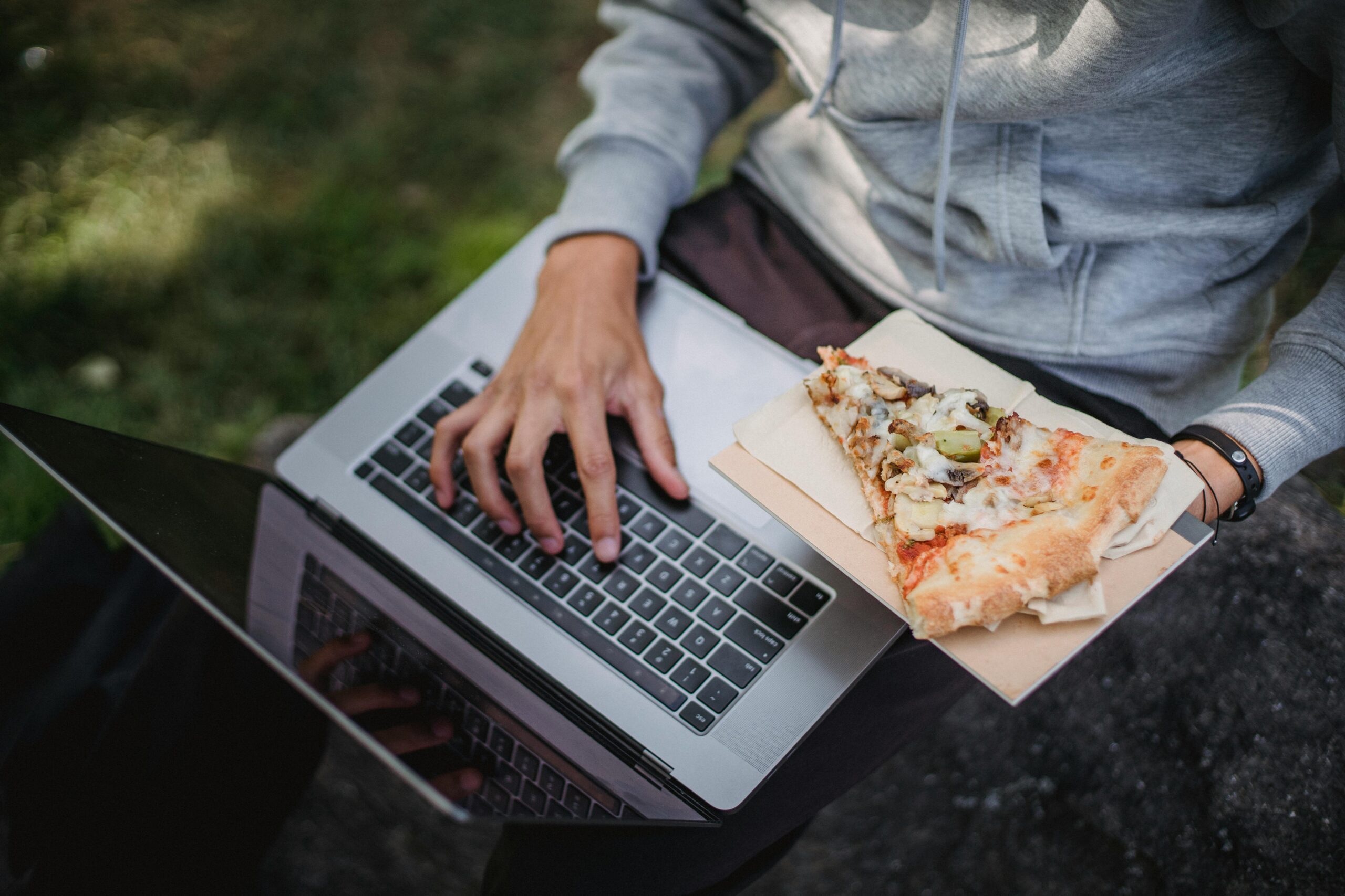 Person wearing a grey hoodie using a laptop outdoors while holding a slice of pizza on a napkin.