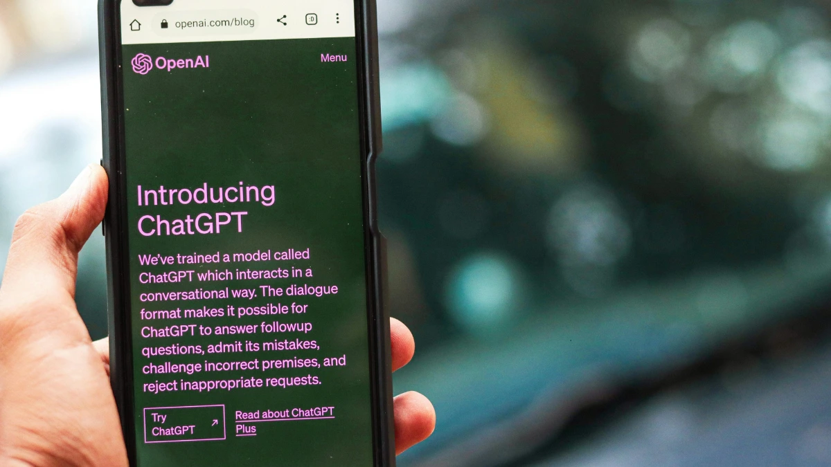 Close-up of a smartphone screen displaying the OpenAI website introducing ChatGPT, with text describing its conversational AI capabilities, held in a person’s hand.