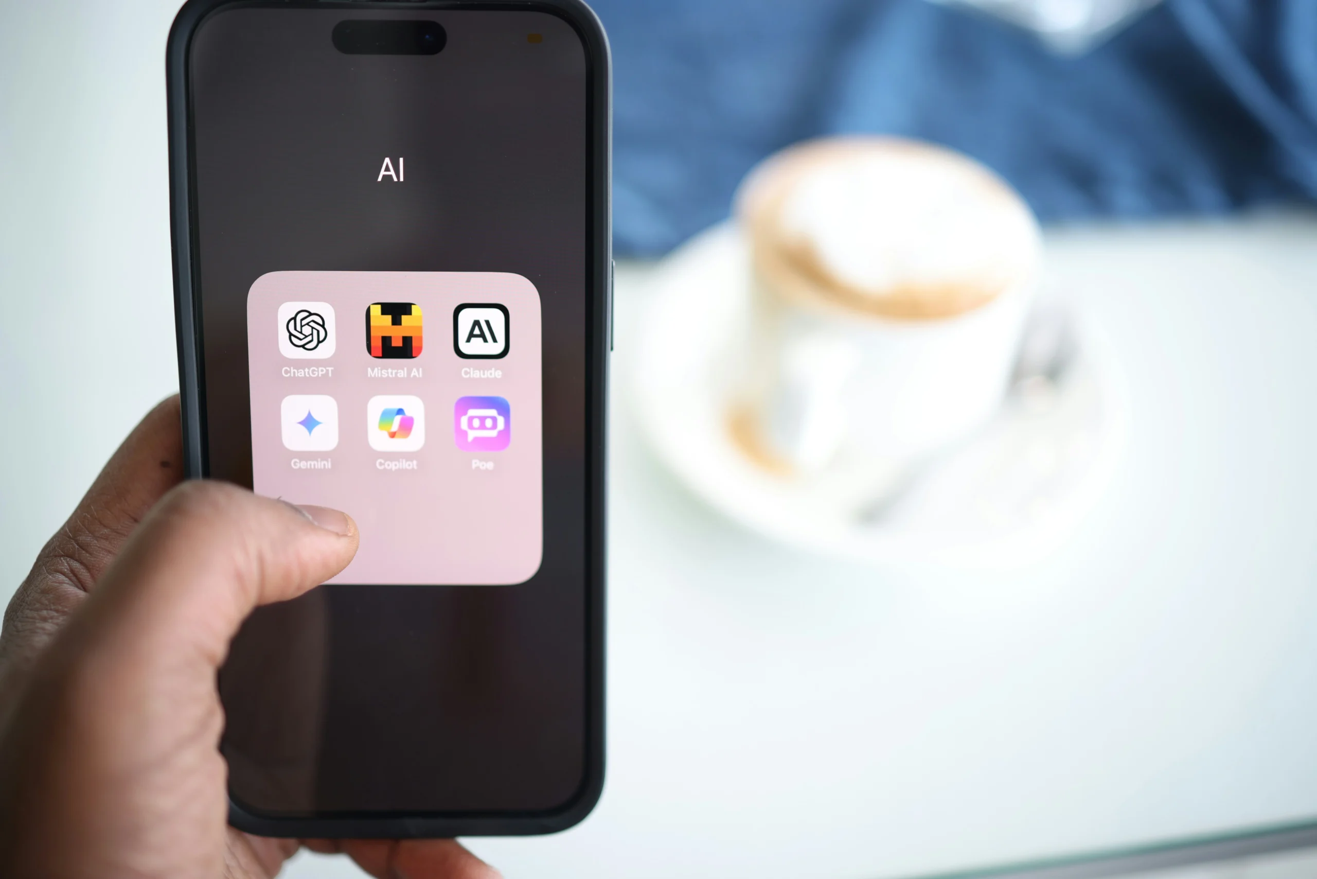 Smartphone screen showing a folder labeled “AI” with popular artificial intelligence apps including ChatGPT, Mistral AI, Claude, Gemini, Copilot, and Poe, held in a person’s hand with a coffee cup blurred in the background.